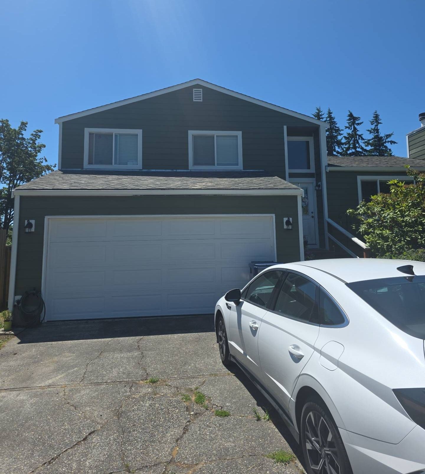 A two-story green house with a white garage door and a white car in the driveway on a sunny day.