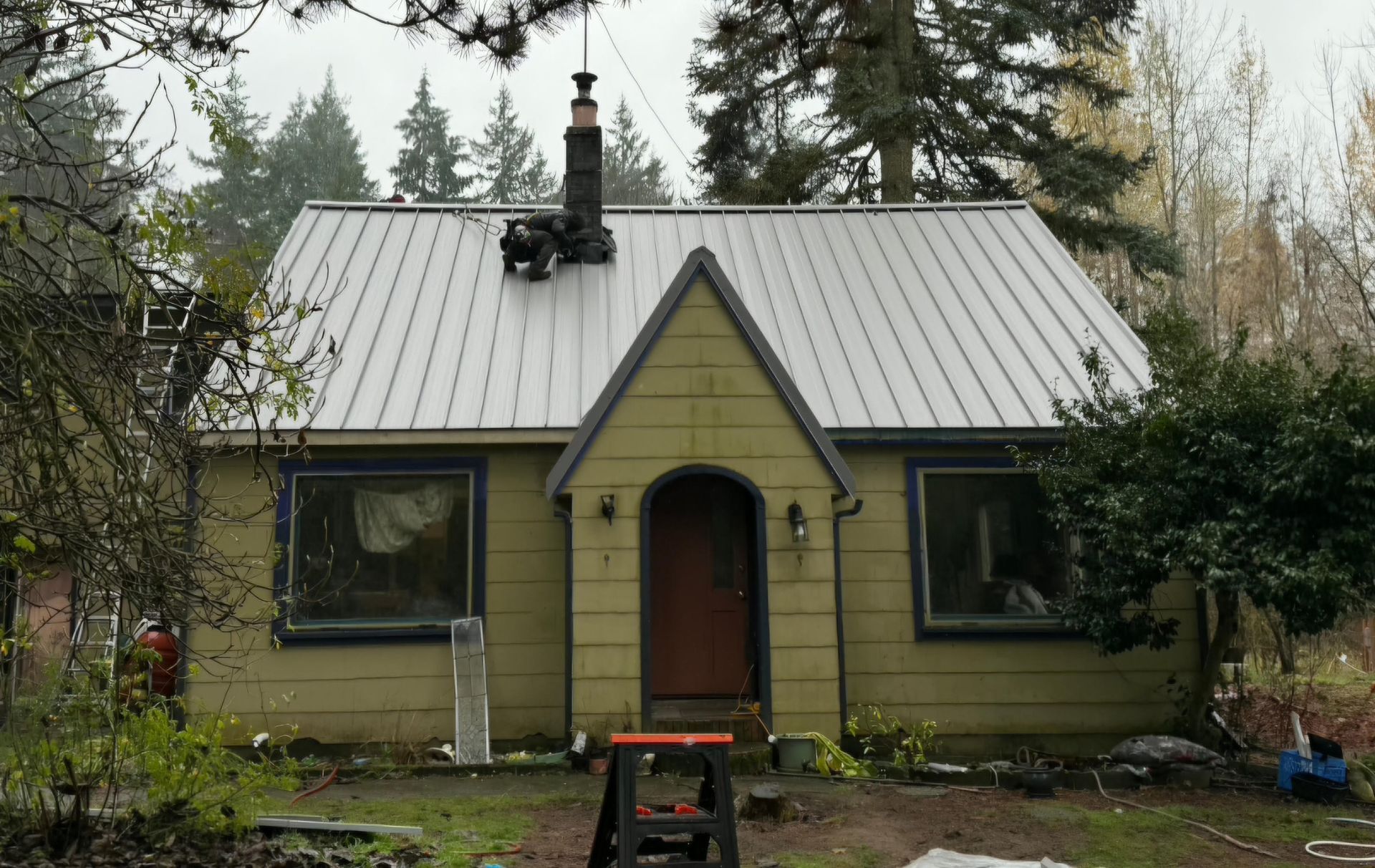 Yellow house with metal roof, chimney, two windows, and dark trim, in a wooded setting.