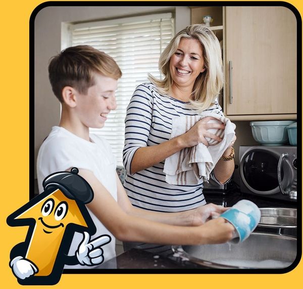 Woman drying dishes while a boy washes in the kitchen sink. Both smiling.
