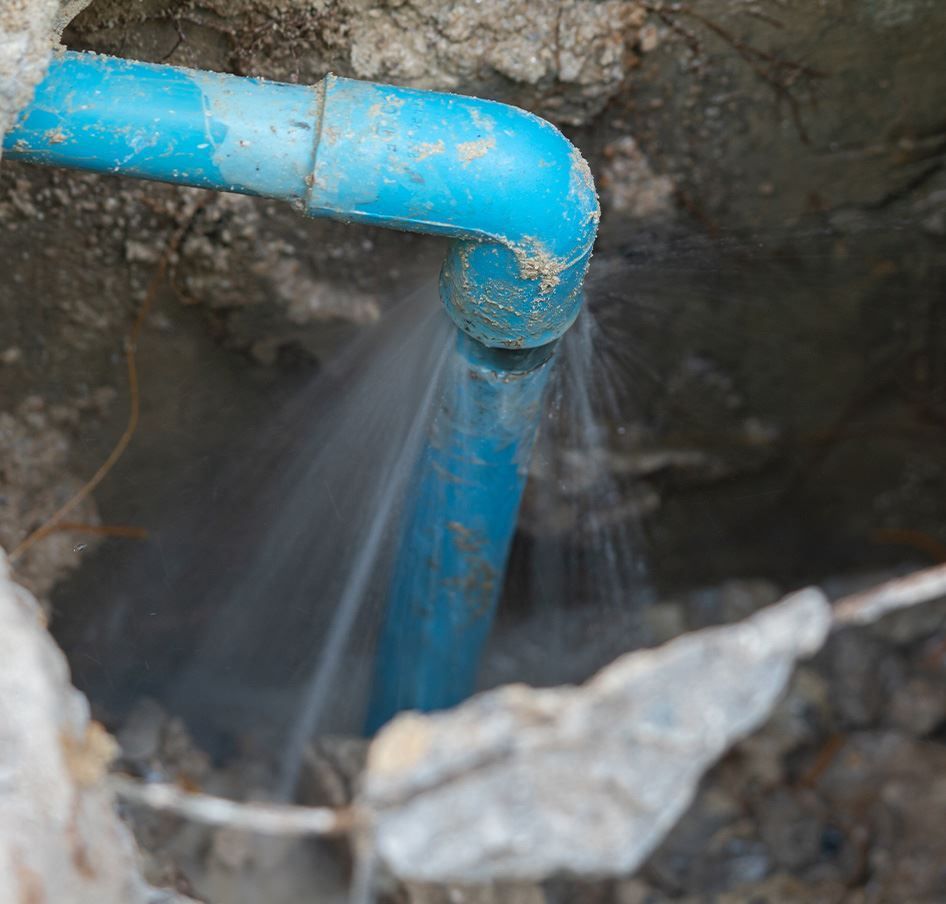 Leaking blue PVC pipe in a dirt-filled trench, water spraying out.