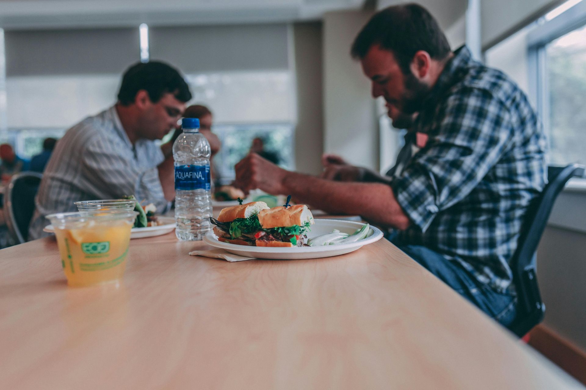 Two people eating at a table in a cafeteria. One eats a sandwich, the other looks down at food.