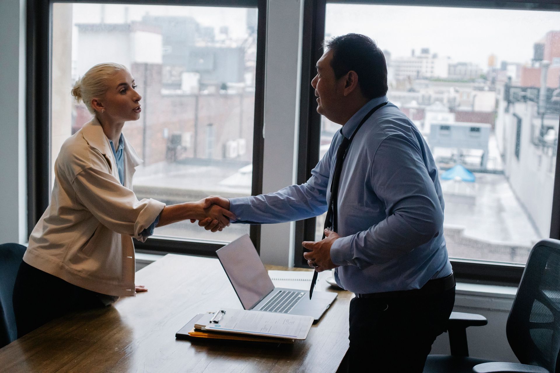 Two coworkers shaking hands across a desk in a bright office with a laptop and city view.