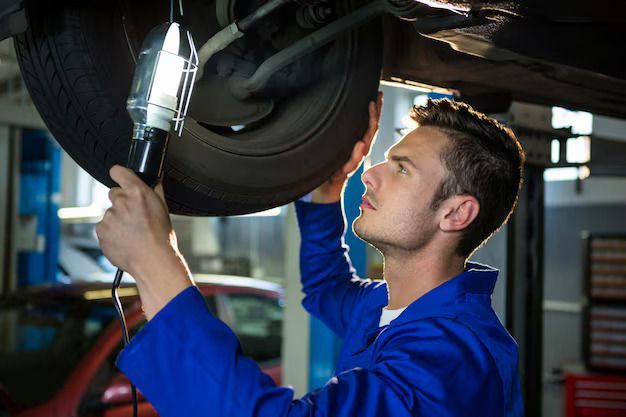 Mechanic in blue overalls inspects car undercarriage with a light in a garage.