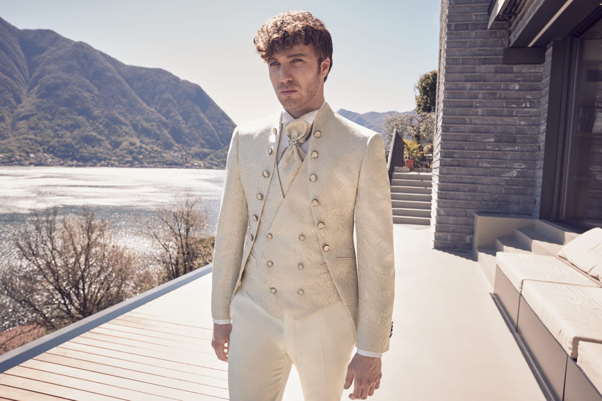 Uomo in abito bianco su una terrazza con lago e montagne sullo sfondo.