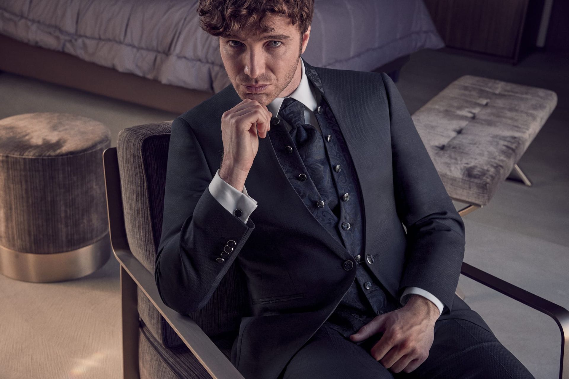 Uomo in abito scuro, gilet e cravatta, seduto su una sedia moderna, ambiente da camera da letto.