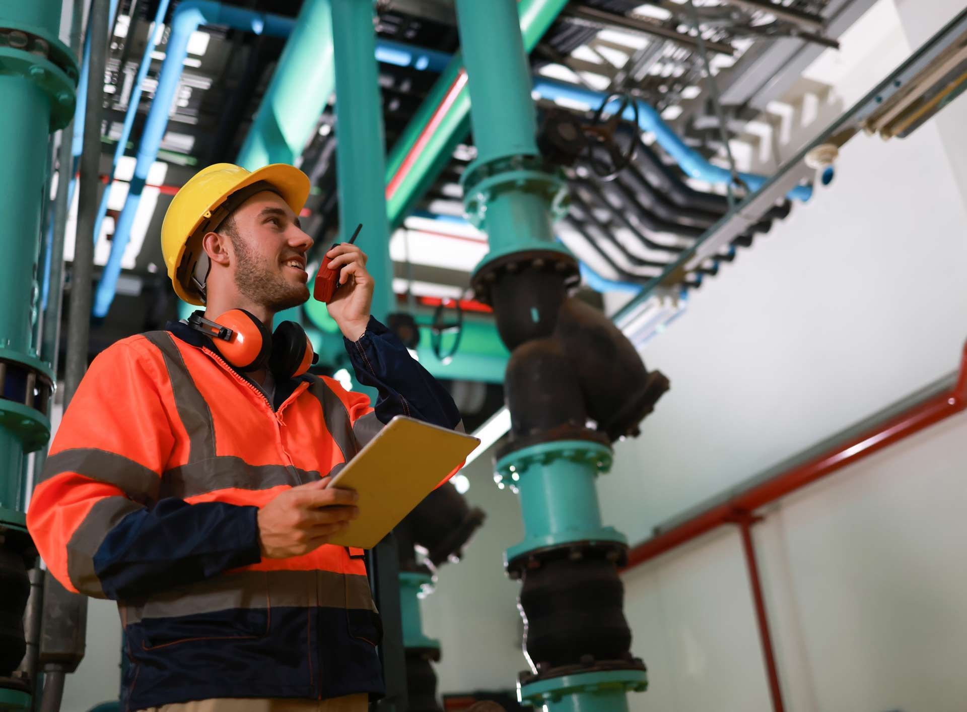 A male plumber wearing an orange safety jacket is inspecting the piping.