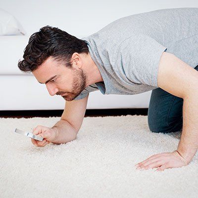 A man is kneeling on the floor looking at something with a magnifying glass.