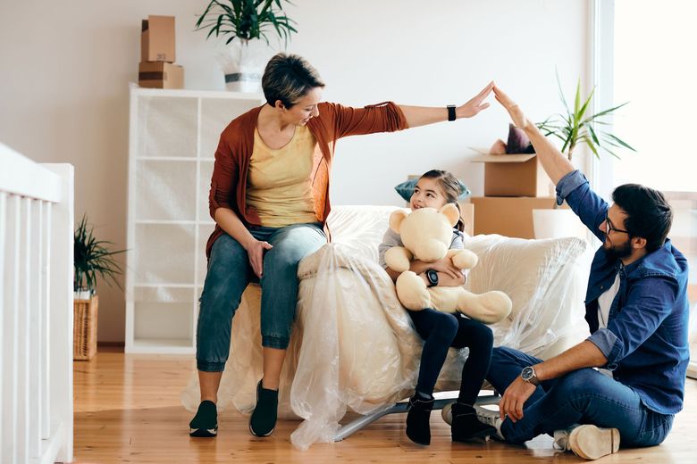 La famille, installée dans sa nouvelle maison, confectionne un toit de fortune au-dessus de sa fille assise sur le canapé recouvert ; des cartons de déménagement se trouvent à proximité.