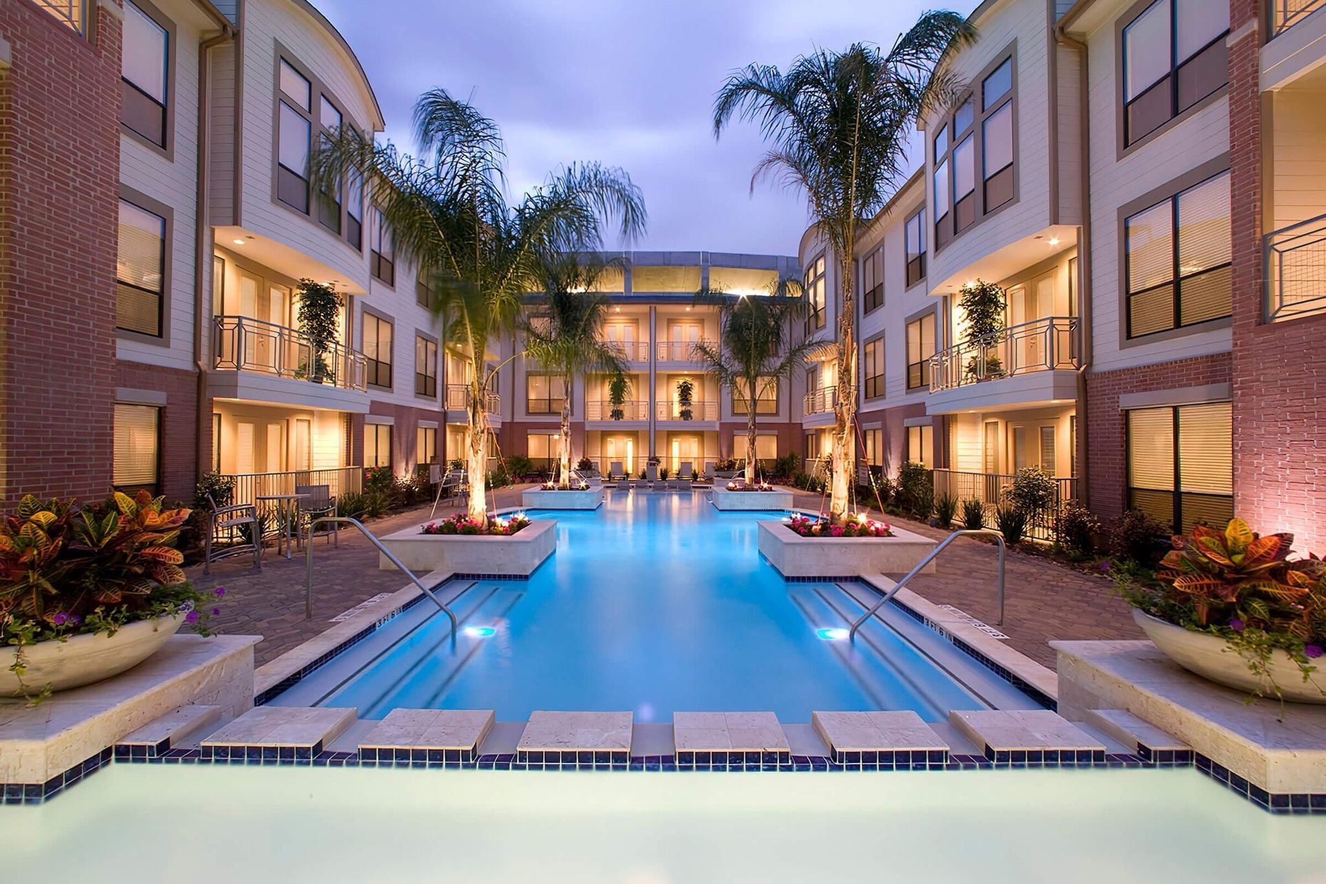 Apartment complex courtyard with pool and palm trees, lit up at dusk.