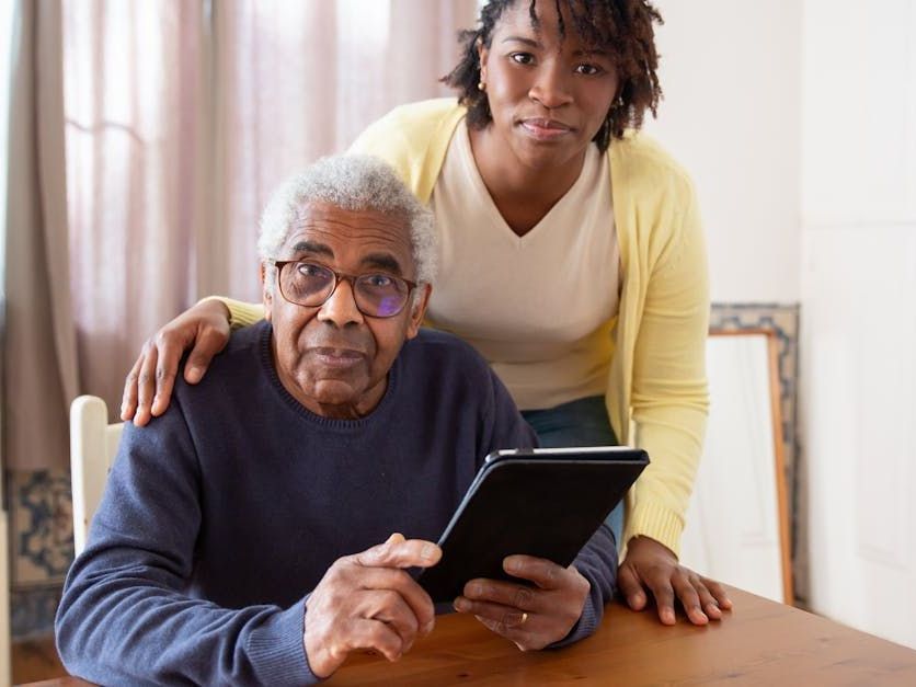 An elderly man is sitting at a table using a tablet computer while a woman stands behind him.