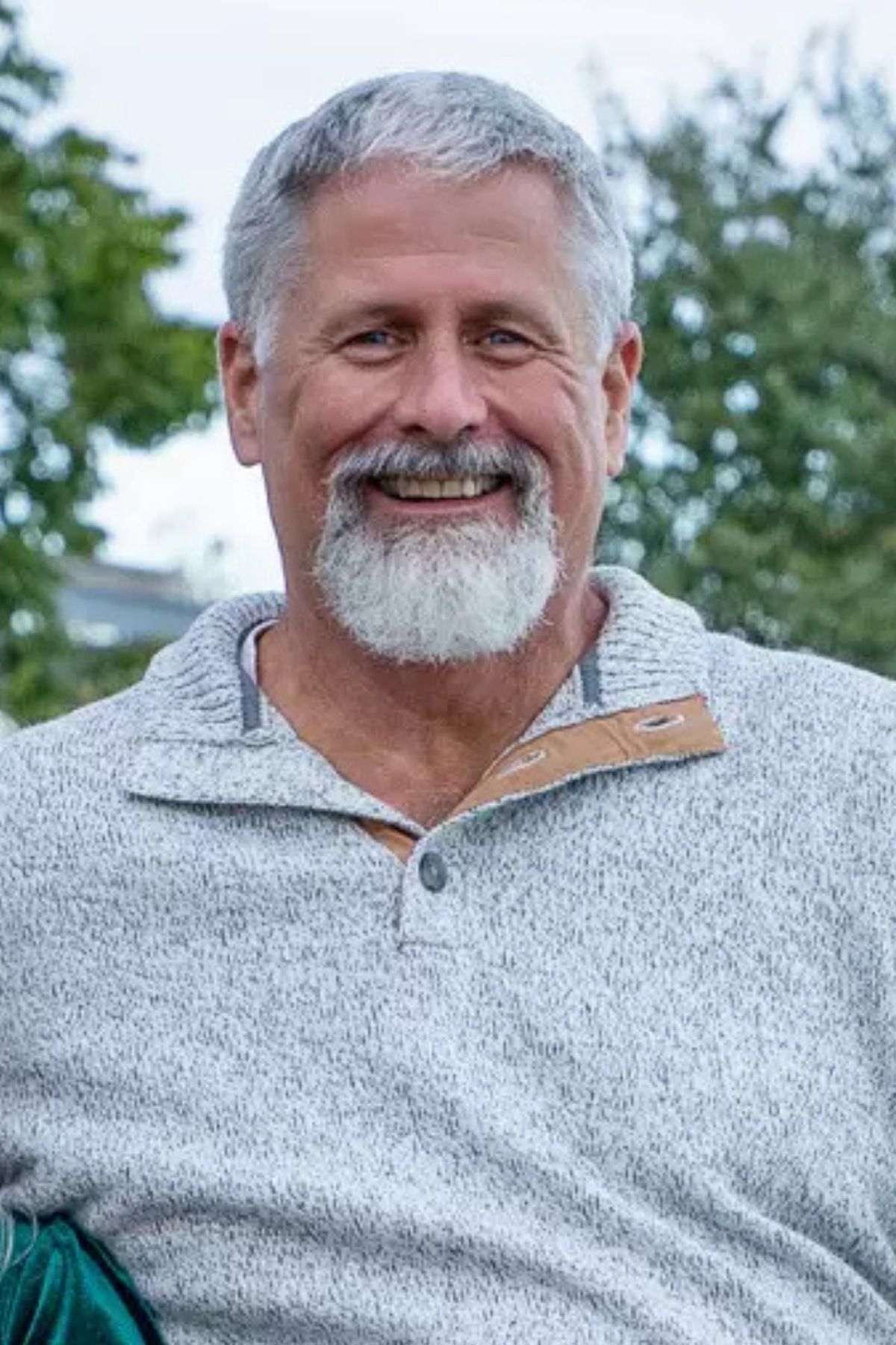 Smiling person with gray hair and a beard, wearing a gray, textured collared sweater outdoors.