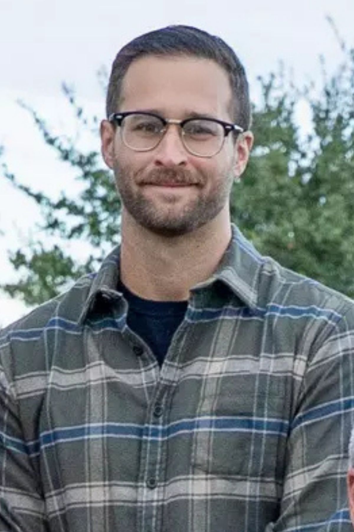 A smiling person with a beard and glasses wears a green and blue plaid flannel shirt against an outdoor backdrop.