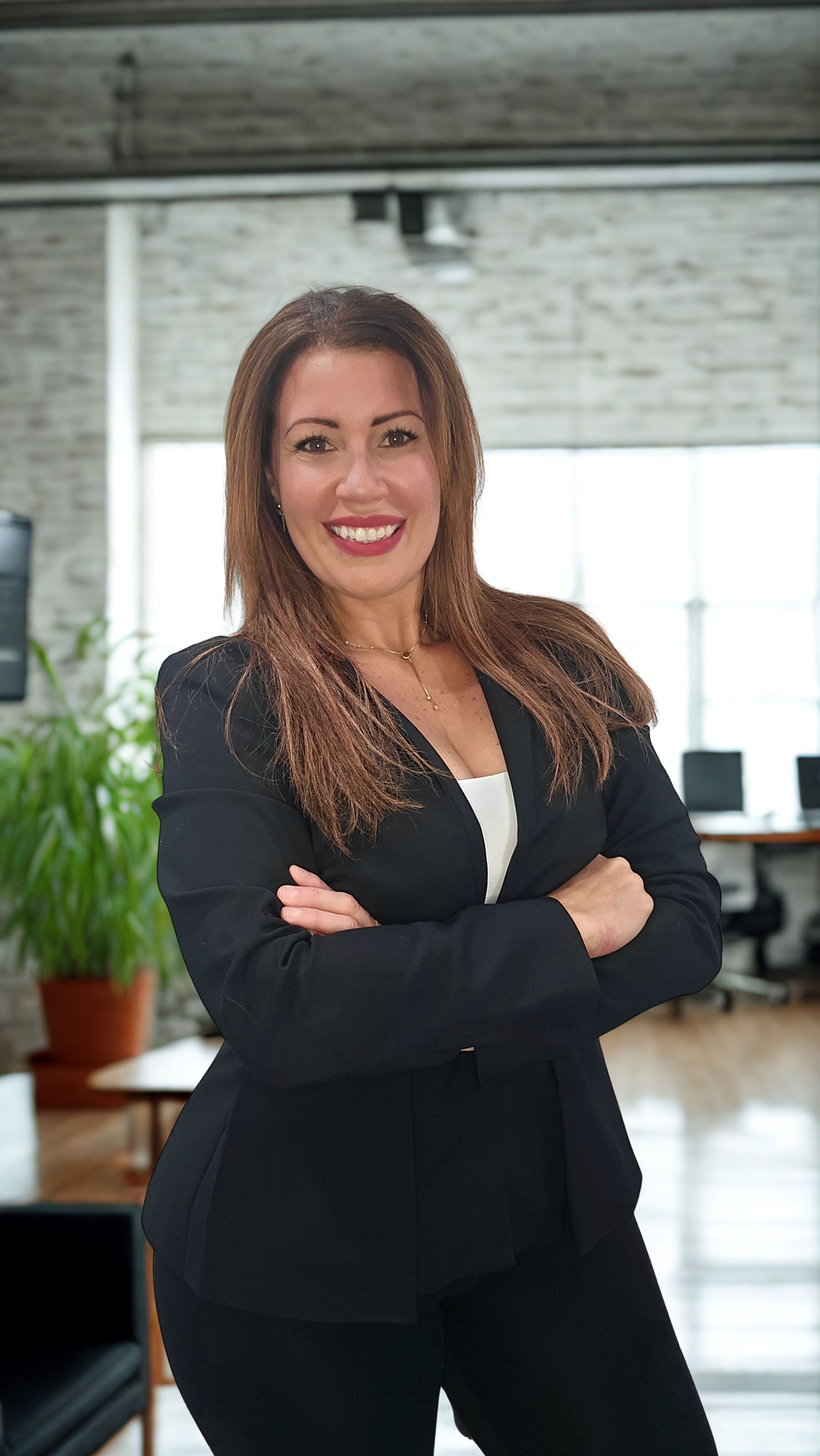 Woman in a black blazer with arms crossed, smiling; office setting with brick wall.