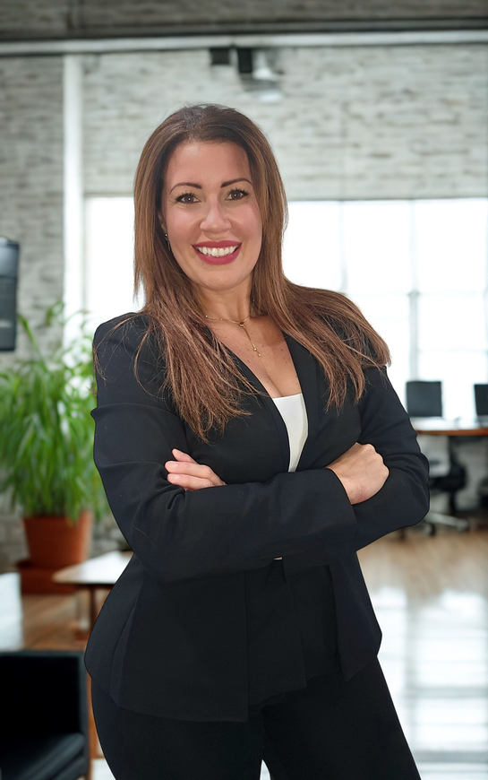 Woman in a black blazer with arms crossed smiling in a modern office setting.