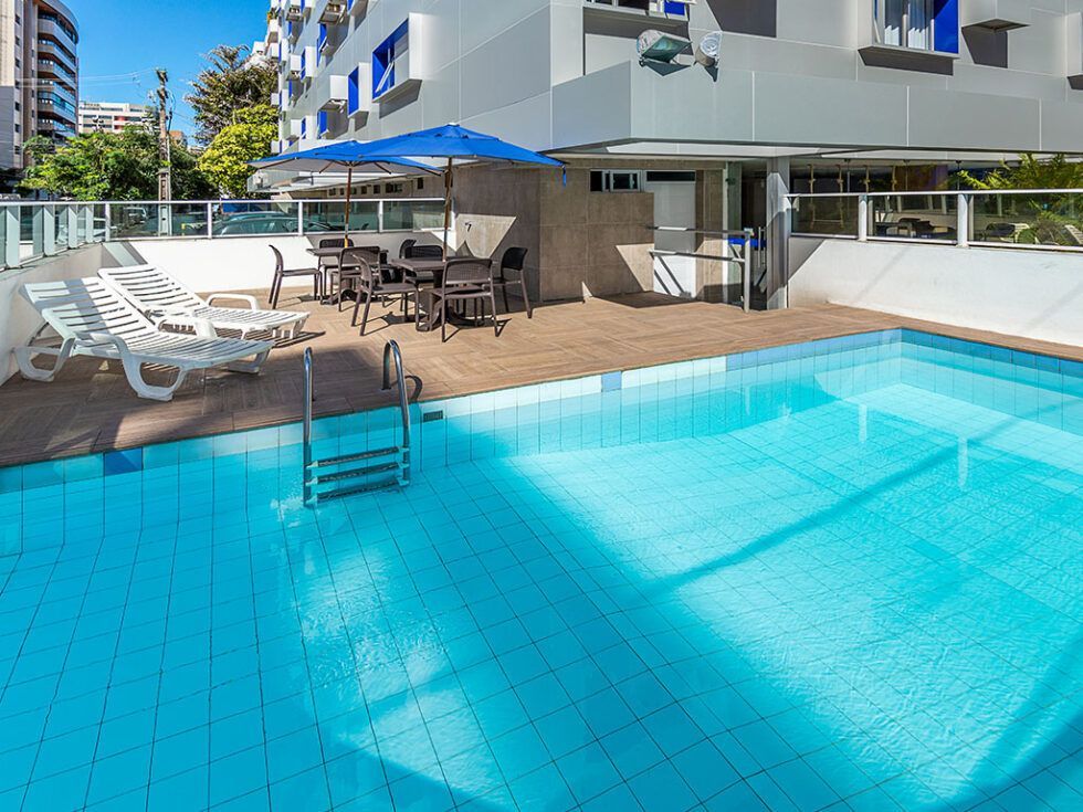 Deck de madeira com mesa, cadeiras e redes sob guarda-sol, palmeiras e céu azul.