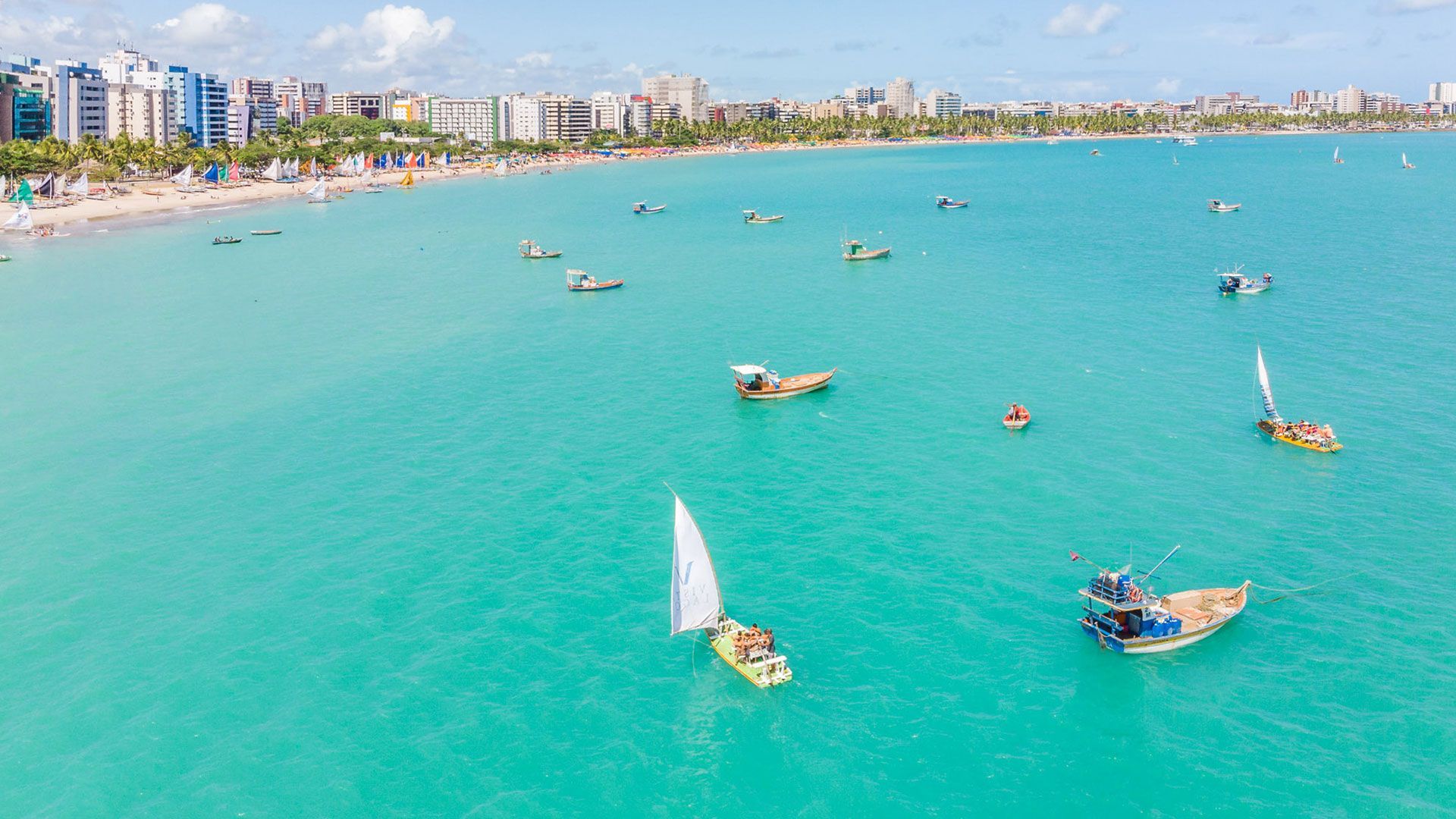 Vista aérea de uma praia azul-turquesa com barcos navegando. Prédios margeiam a costa sob um céu azul claro.