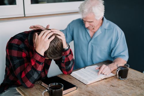Man comforts another over a Bible at a table. One man has head in hands, the other touches his head.