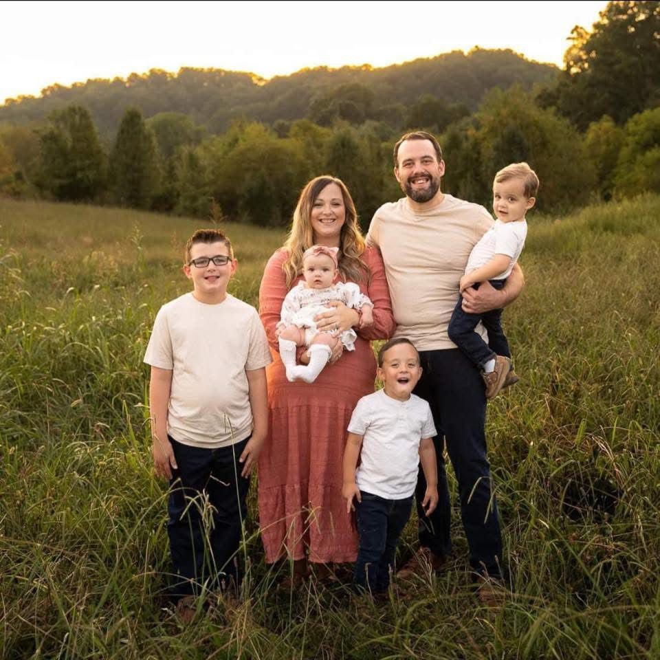 Family of five in a grassy field; mother in a rust dress holding a baby, father with a child, two boys smiling.
