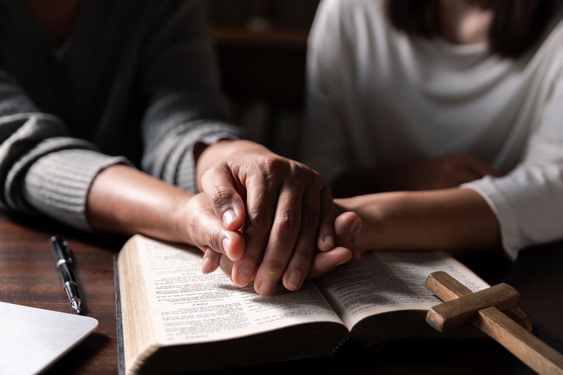 Two people holding hands over an open Bible with a wooden cross, lit low.