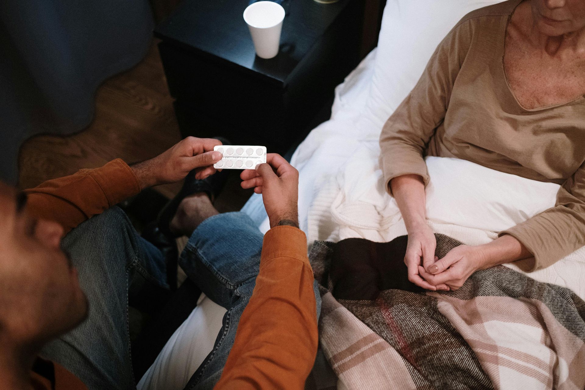 Person giving medication to someone in bed; bedside table, drink, blanket visible.