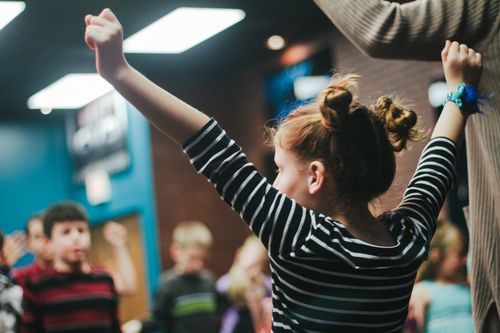 Girl with red hair in a striped shirt raises arms in a room with other children and an adult.