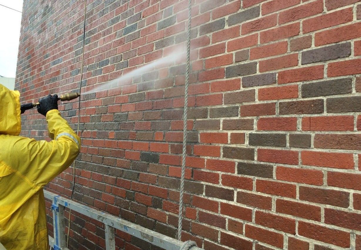 A man in a yellow raincoat is cleaning a brick wall with a high pressure washer.