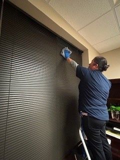 A woman is standing on a ladder cleaning blinds.
