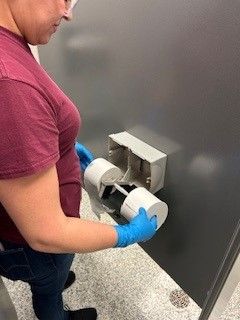 A woman is taking a roll of toilet paper out of a dispenser in a bathroom.