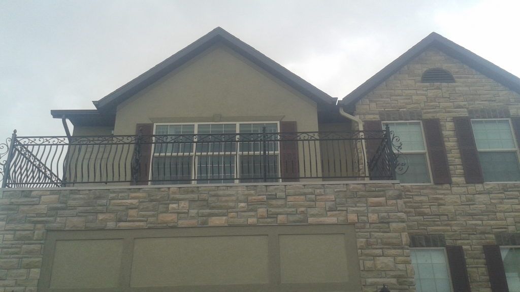 Two-story house with stone facade, balcony with black railing, tan stucco, and brown shutters.