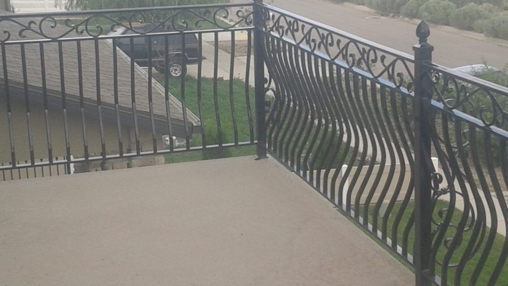 Black metal balcony railing with wavy and straight vertical bars, decorative top, overlooking a yard.
