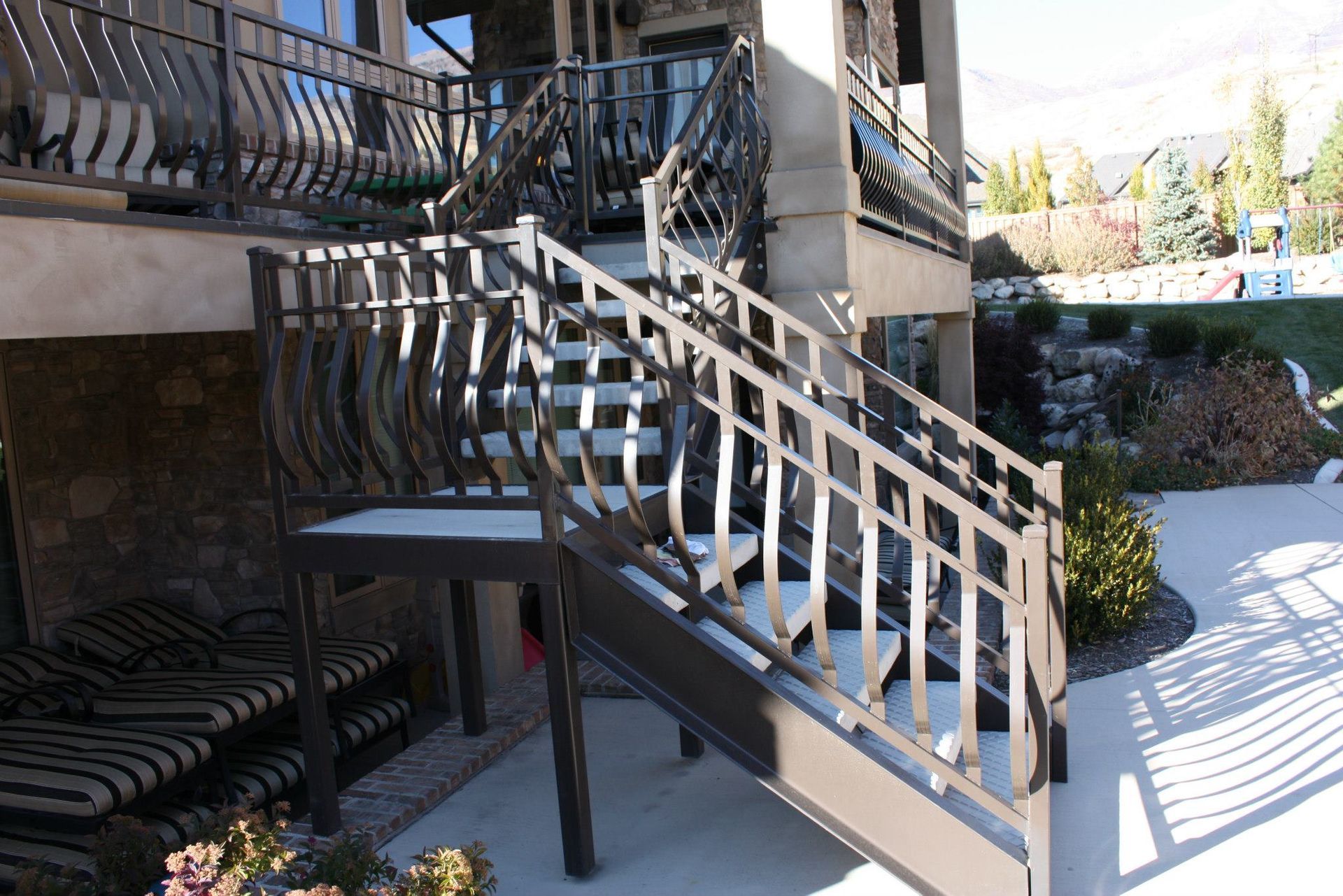 Outdoor staircase with decorative metal railing leading to a deck, adjacent to a house with a stone facade.
