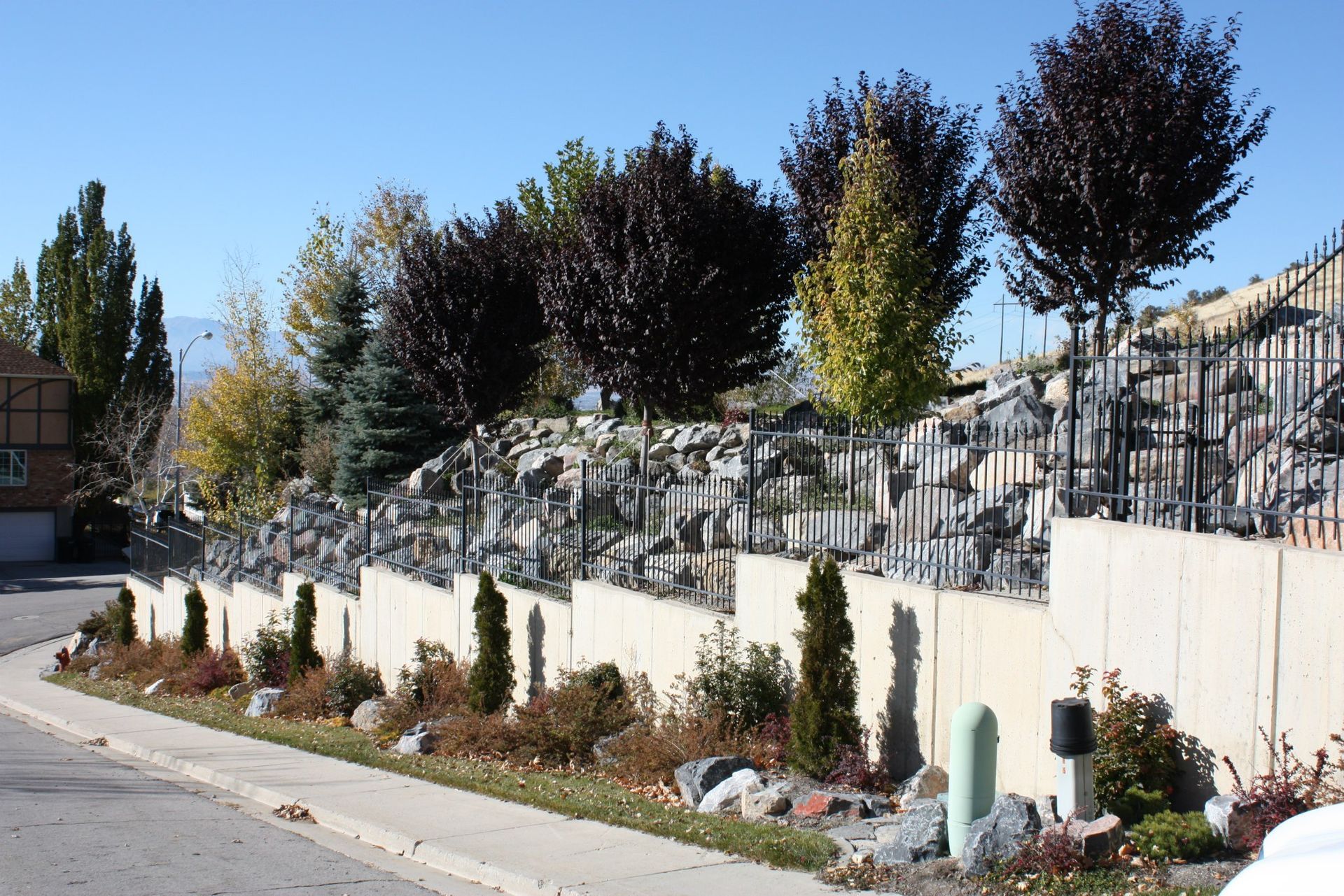 Stone retaining wall with trees and shrubs along a curved street under a blue sky.