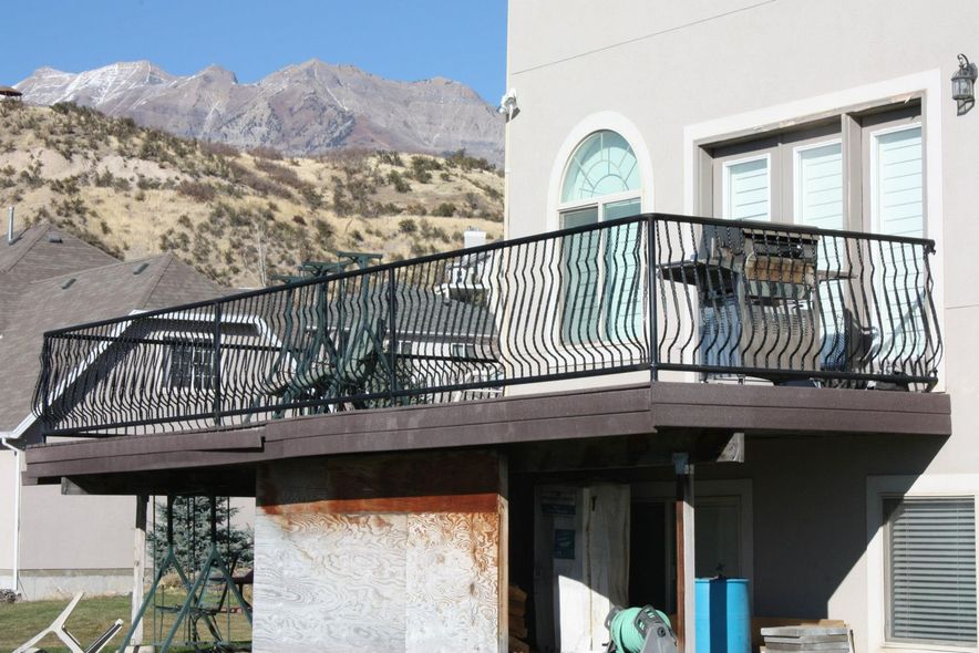 Black metal railing on a deck with mountain backdrop. House with white exterior and chairs on deck.