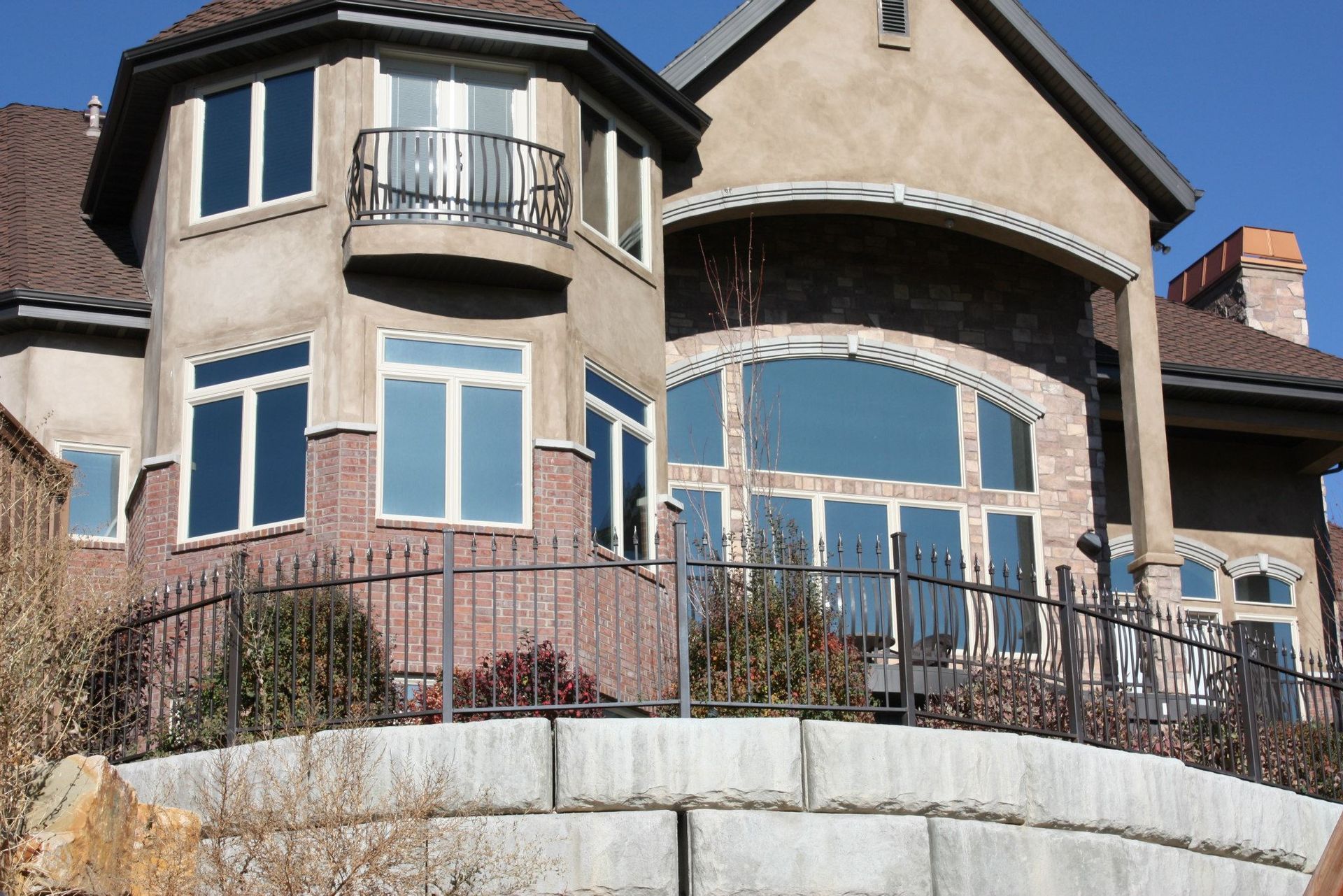 Two-story house with brick and stucco exterior, dark roof, and large arched windows, with a wrought iron fence in front.