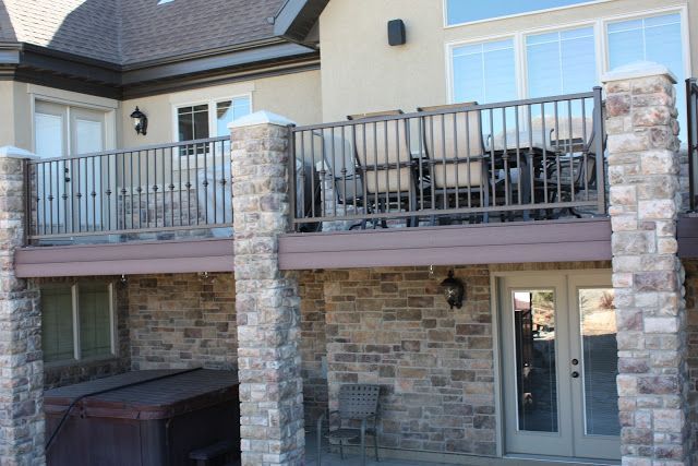 Two-story house with stone columns, deck with metal railing, chairs and table, and a hot tub below.