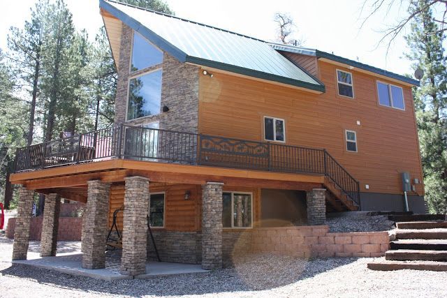Two-story wooden house with stone accents, a large deck, and metal roof, set among trees.