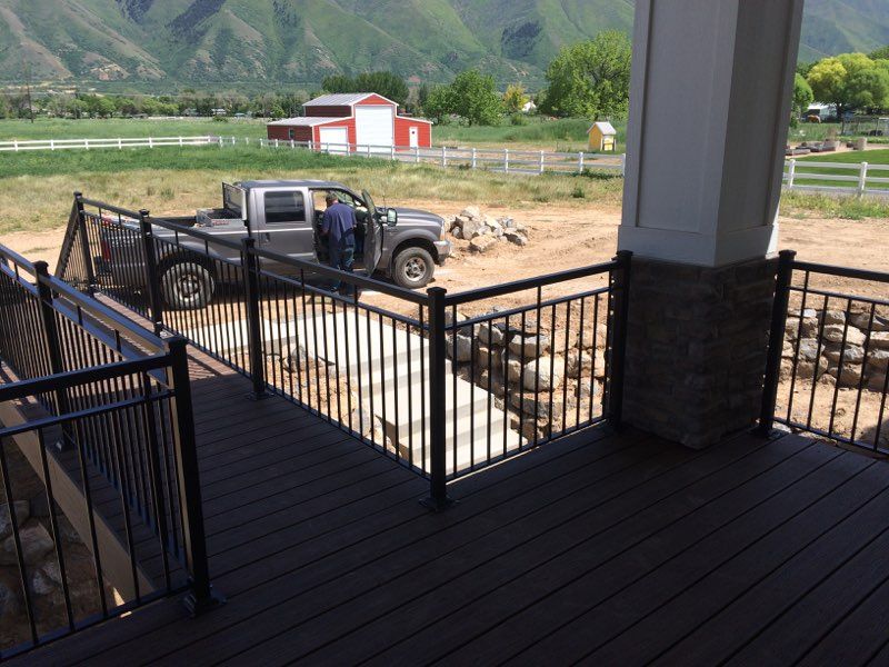 Black railing on a deck; a truck with a person is parked near a dirt area, with a red barn in the background.
