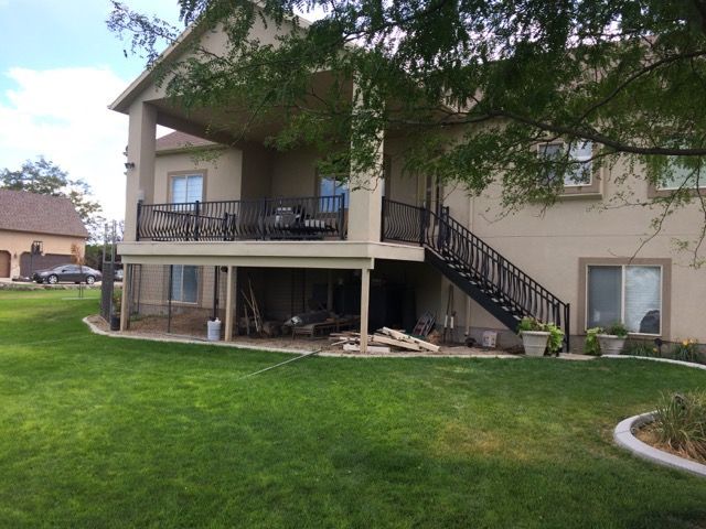 Back of a two-story beige house with a covered deck, black railings, and a staircase leading down to a green lawn.