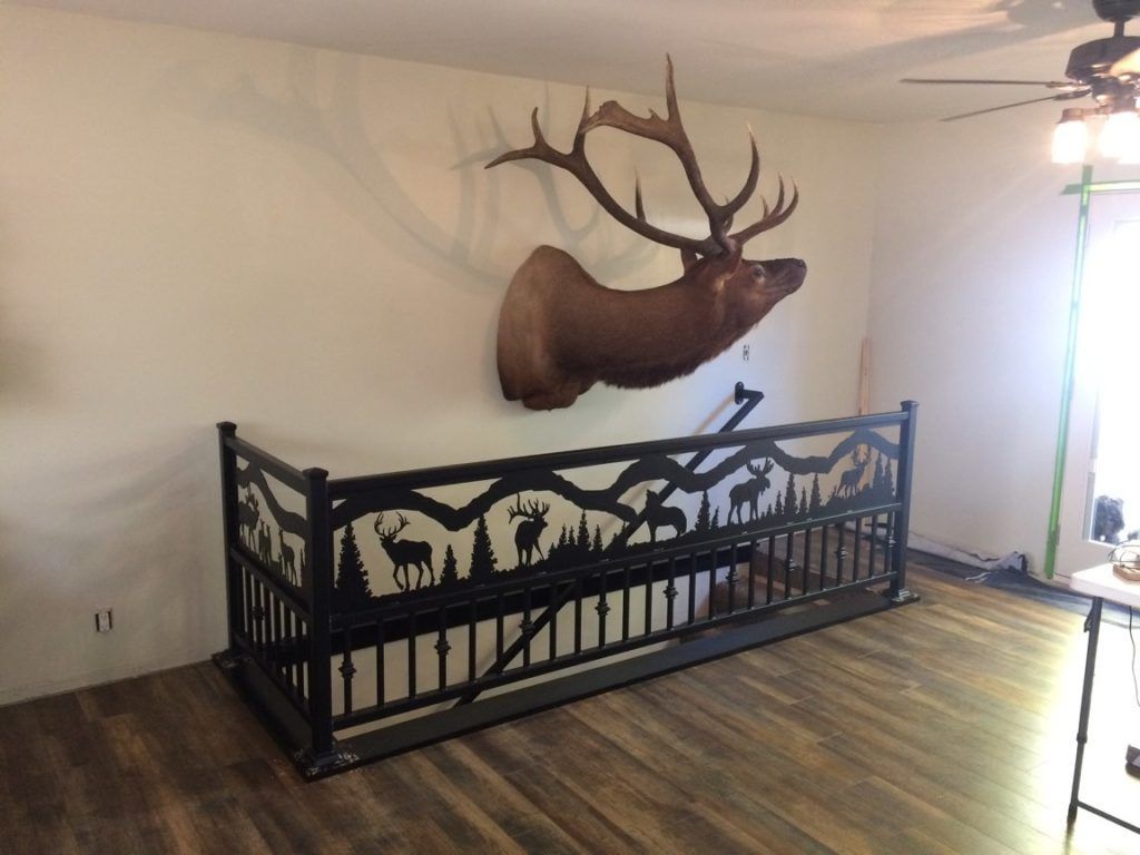 An elk head mount above a black metal railing with forest silhouette designs; a room with wood flooring.