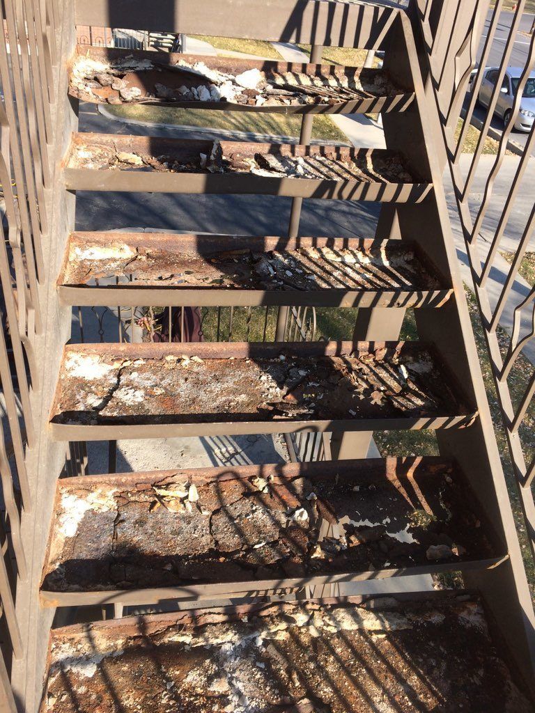 Rusting, weathered metal fire escape stairs. Some treads have missing or damaged sections, casting shadows.
