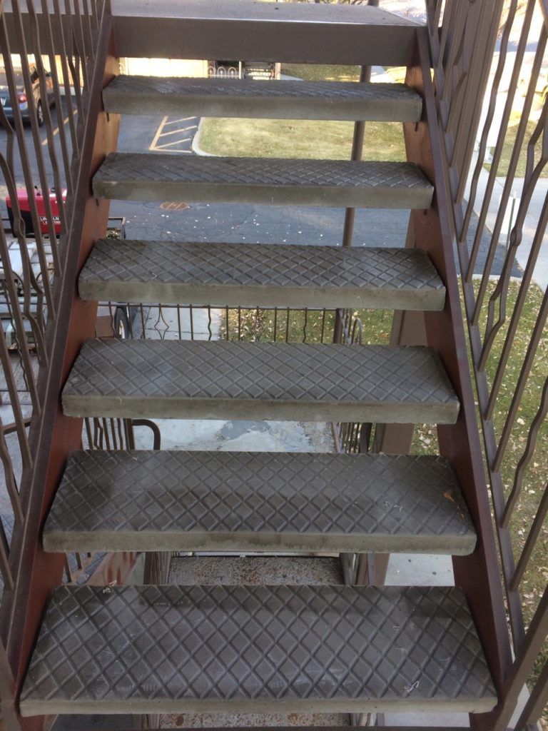 Metal staircase, descending from above. Diamond plate steps and rusty railings are visible, outdoors.