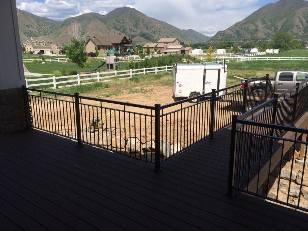 Black metal railing on a deck overlooks a yard with a white fence, trailer, and distant mountains.