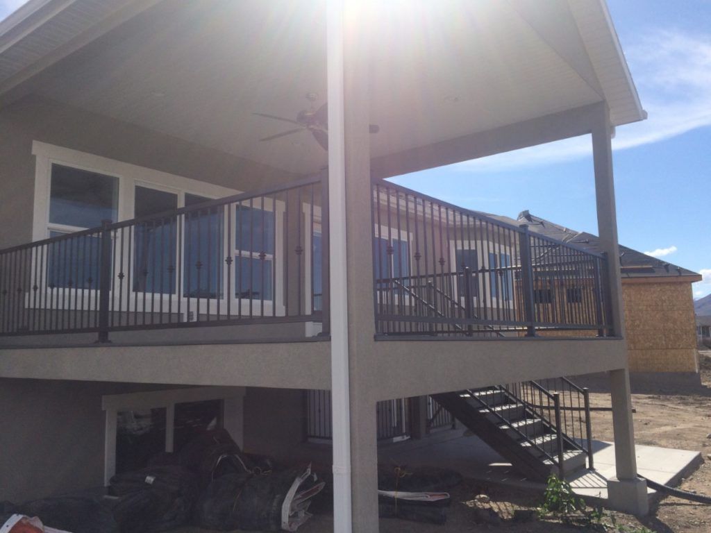 Covered outdoor patio with black railing, stairs, and white trim against a light-colored stucco building under a sunny sky.