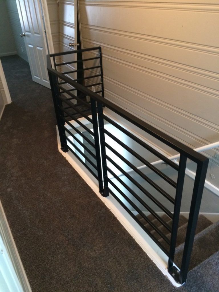 Black metal railing on a staircase with dark gray carpet, against a light-colored paneled wall.