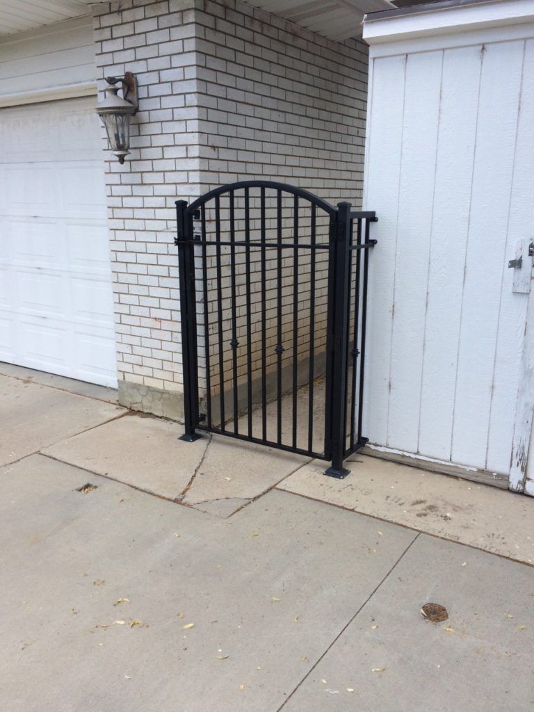Black metal gate between a white brick building and a white wooden fence.