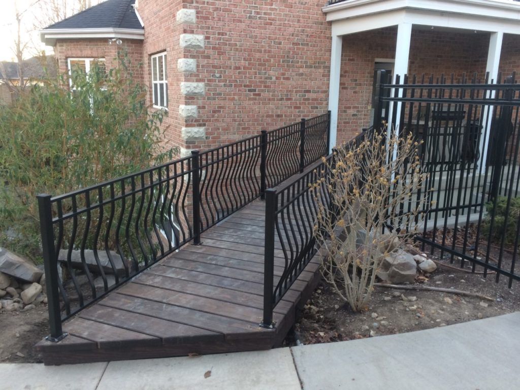 Black metal bridge with decorative railings, over a small ditch next to a brick building.