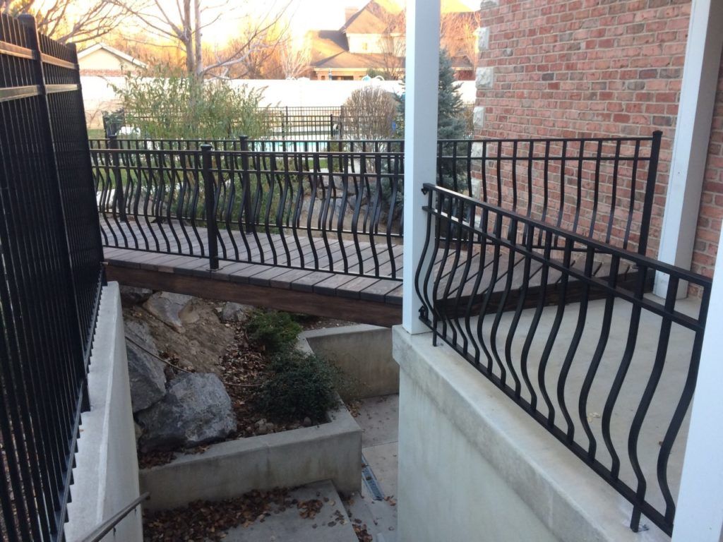 Black metal railings enclose a porch and a walkway. A brick building and landscaping are in the background.