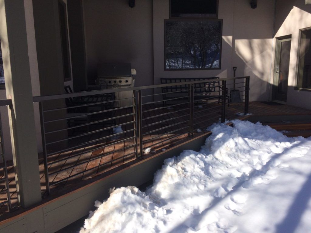 Outdoor deck with metal railing, snow piled along the edge, and a grill in the background.
