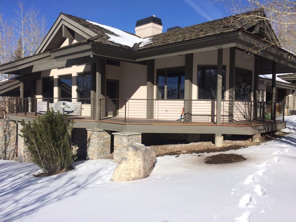 A house with a wraparound deck on a snow-covered landscape. Brown roof, tan siding, black windows.