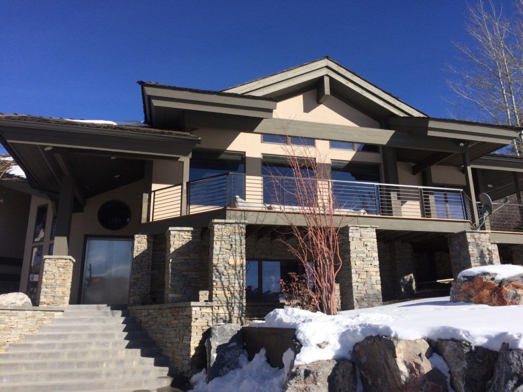Two-story house with stone columns, steps, and snow on the ground under a blue sky.